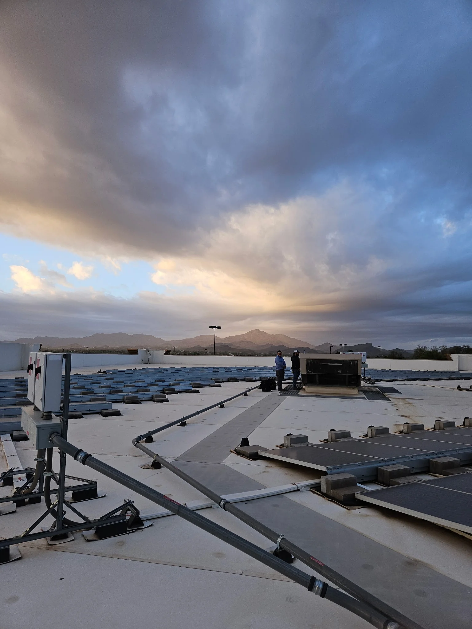 Rooftop solar panel array on white membrane roof with gray racking, conduit runs, and electrical enclosures at dusk