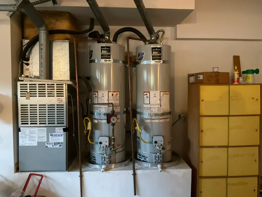 Two stainless steel water heaters with copper piping, expansion tanks, and furnace unit in utility room