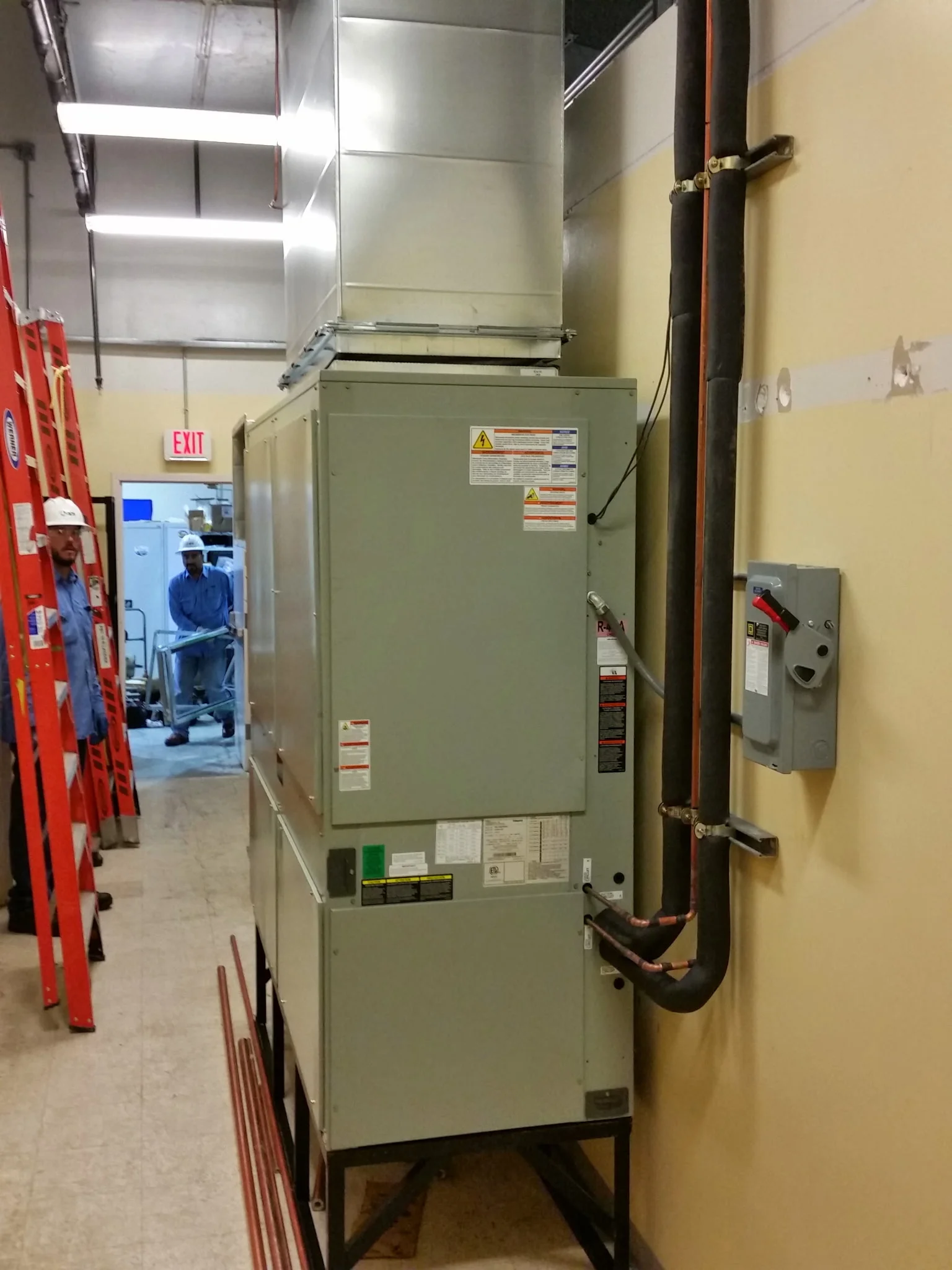 Gray commercial air handler on steel stand with insulated copper refrigerant lines, sheet metal ductwork, and wall-mounted electrical disconnect