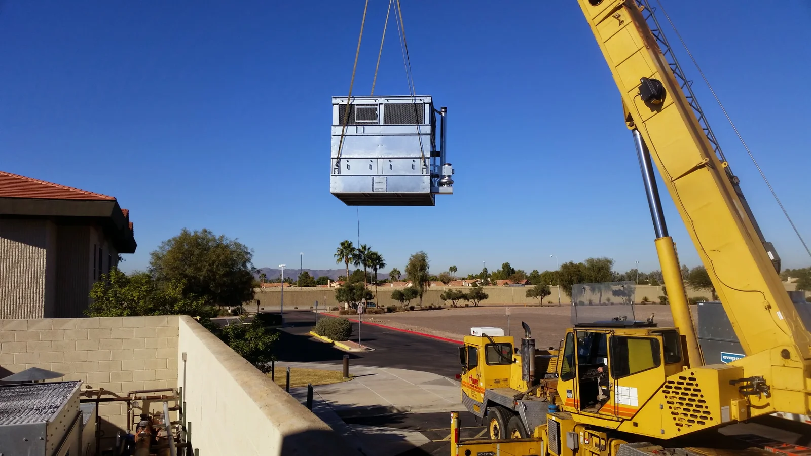 Crane lifting galvanized steel rooftop HVAC unit with lift straps over masonry parapet wall