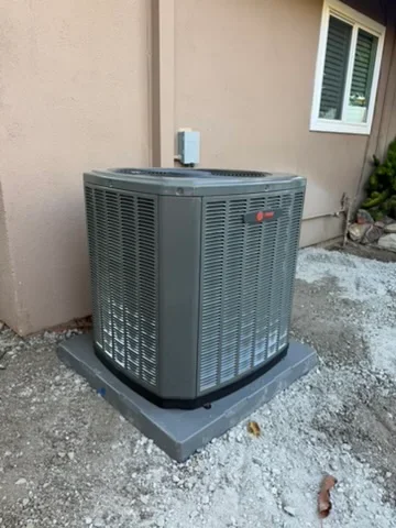 Gray central AC condenser unit installed on concrete pad against tan stucco wall