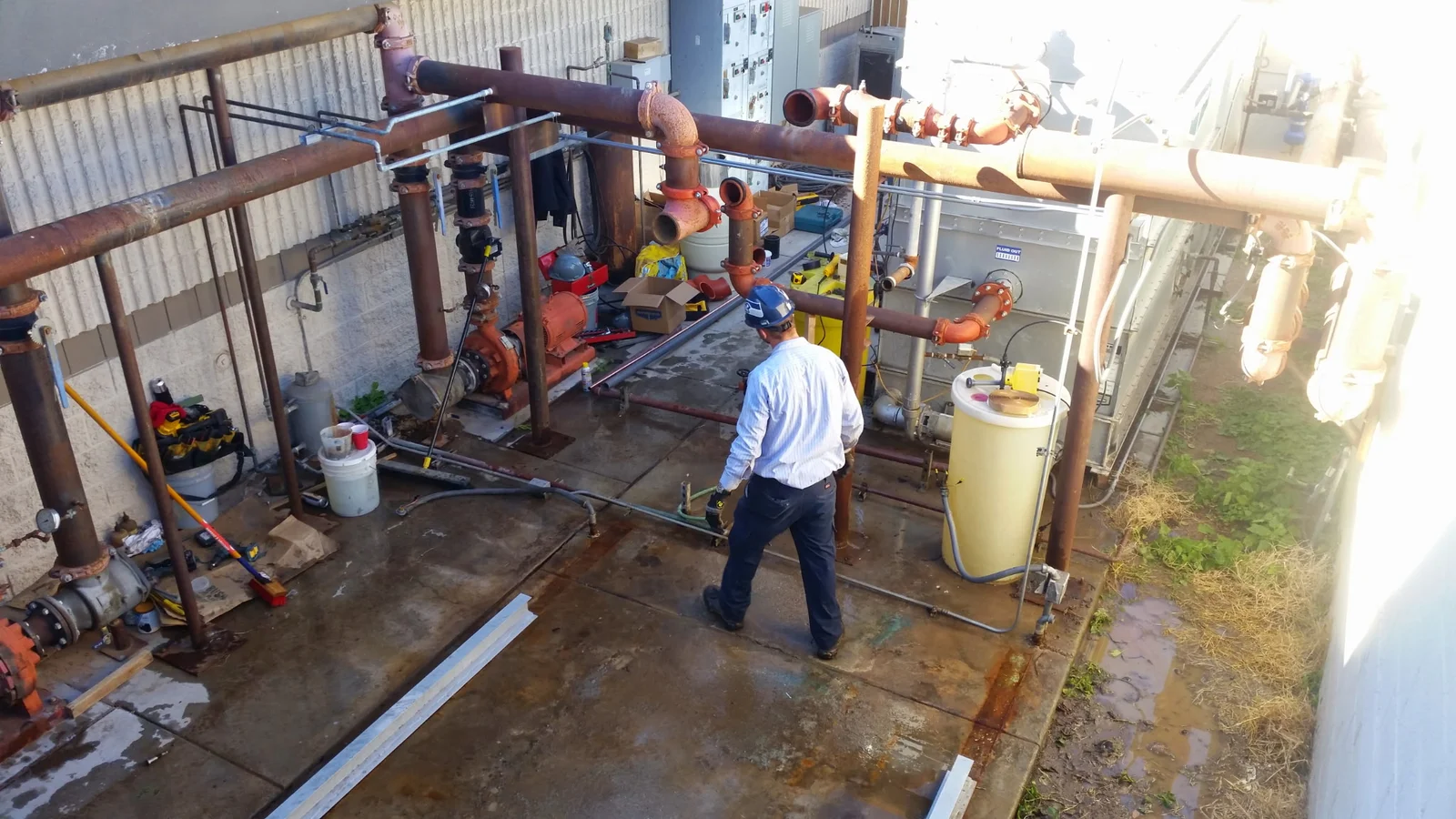 Outdoor steel pump station with rust-colored flanged piping, orange pump casing, and technician inspecting concrete pad